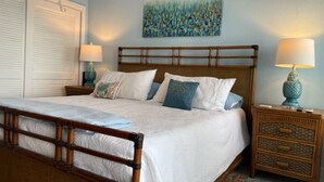 Quarto com cama de madeira, roupa de cama branca, mesa de cabeceira com abajur e um quadro na parede.