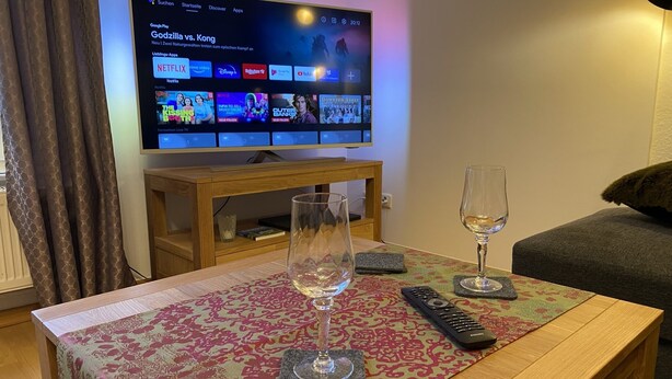 A living room with a flat-screen TV displaying a Netflix interface, a remote control, two wine glasses, and a patterned tablecloth.
