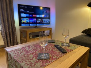 Ein Wohnzimmer mit Fernseher, der einen Film zeigt, einem Tisch mit Fernbedienung und Glas sowie einer Couch.