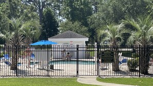 Zona de piscina cercada con sillones de descanso y sombrillas, un edificio pequeño y un cartel que indica los horarios y normas de la piscina.