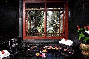 Un baño con pared de azulejos negros, una ventana con marco rojo, una bañera con pétalos de flores y una planta en maceta con flores rojas.