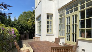 A patio with a wooden table and chairs, surrounded by a white building with large windows. - Large beautiful unusual house with lots of character and art on the walls. (Bournemouth)