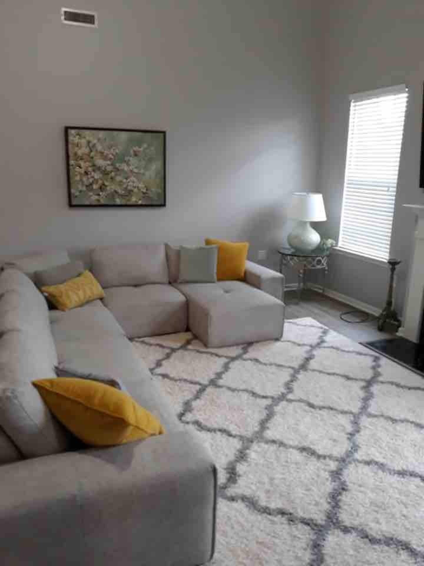 Ein Wohnzimmer mit einem Ecksofa, einem Teppich mit Muster, einer Lampe und einem Fenster mit Jalousien.