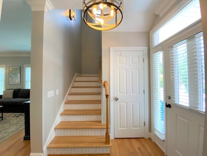 A staircase with patterned carpet leading to a white door, a chandelier above, and a view of a living room with a couch and artwork. - Pink Flamingo | Yacht Basin & Marsh Views | 3 Blocks to Beach | Sleeps 10 | Dog Friendly (Edisto Beach)