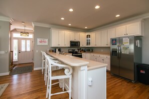 A modern kitchen with white cabinets, a stainless steel refrigerator, and a bar area with white chairs. - Pink Flamingo | Yacht Basin & Marsh Views | 3 Blocks to Beach | Sleeps 10 | Dog Friendly (Edisto Beach)