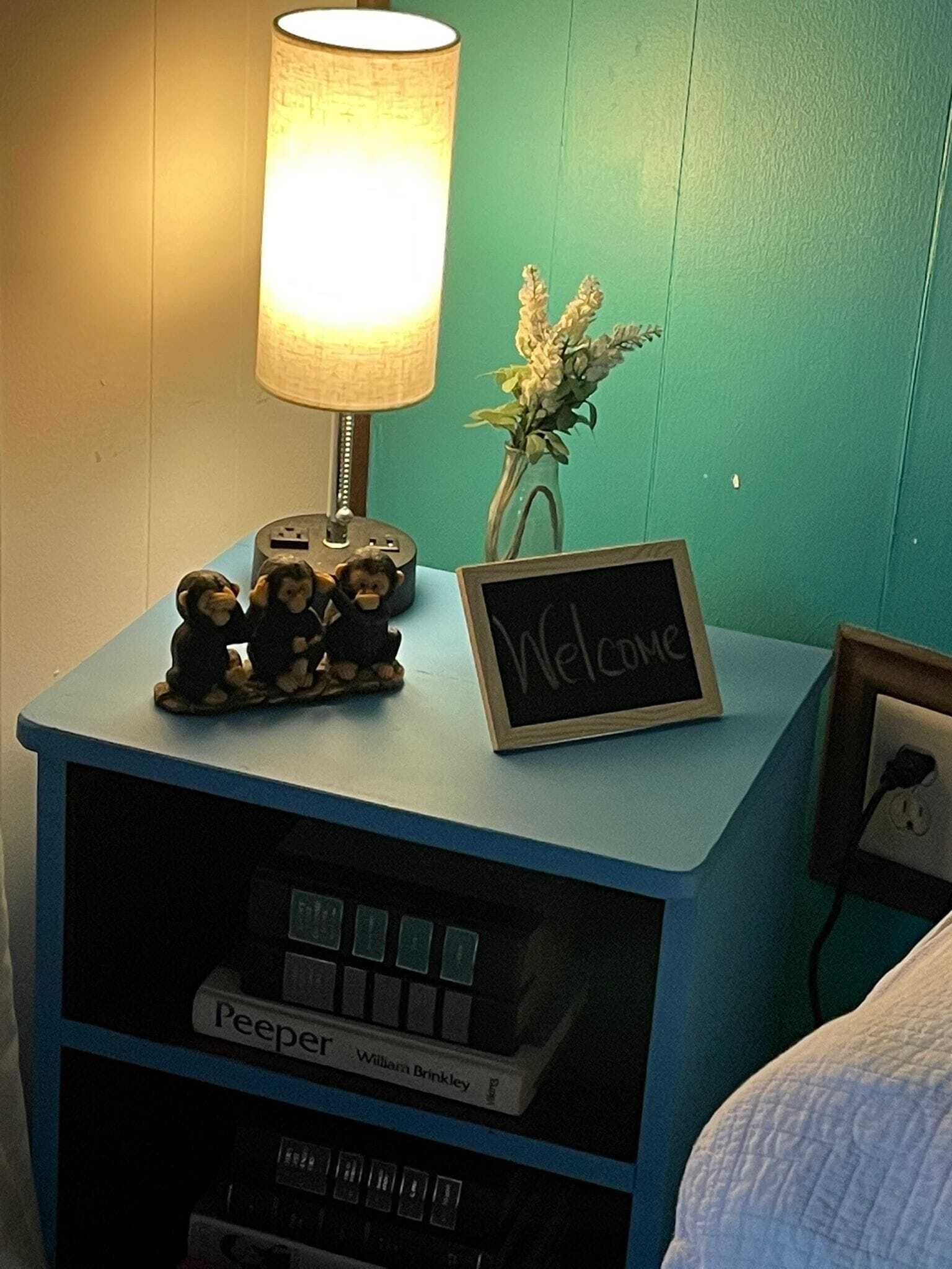 A small bedside table with a lamp, a welcome sign, a small figurine, and a book titled 'Peeper'.