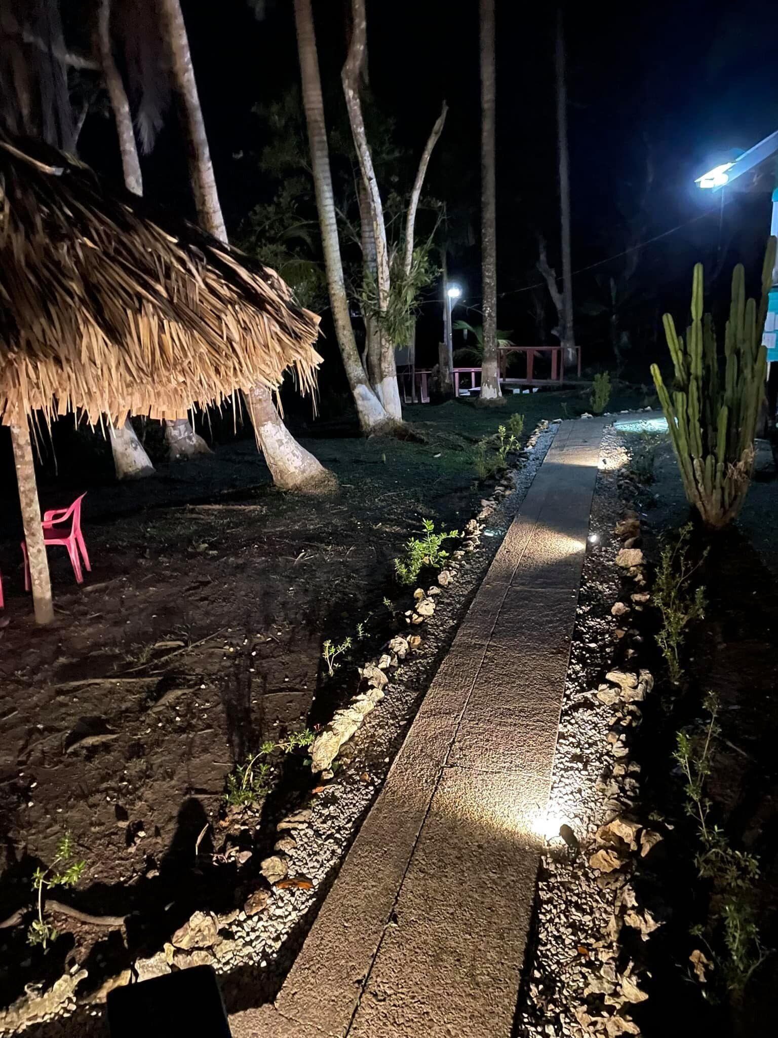 Um caminho com pedras e plantas, que leva a uma construção de palha com uma cadeira rosa.