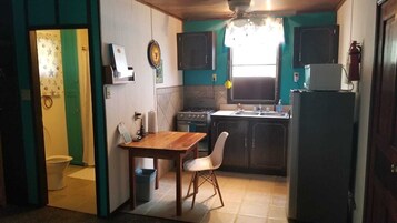 Una cocina compacta con estufa, lavamanos y refrigerador. Hay una mesa de comedor con sillas y un pequeño bote de basura.