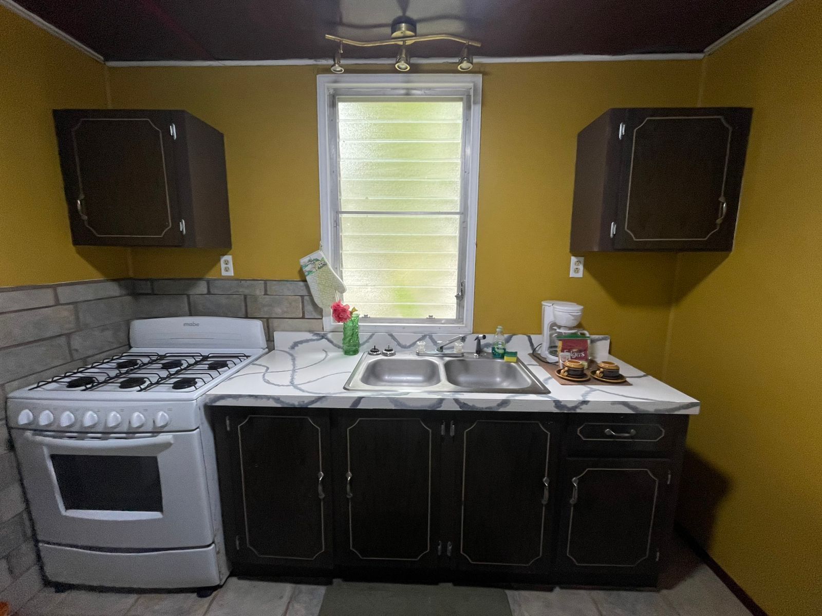 A compact kitchen with a gas stove, oven, and a sink. There are cabinets above and below the countertop, and a window above the sink.