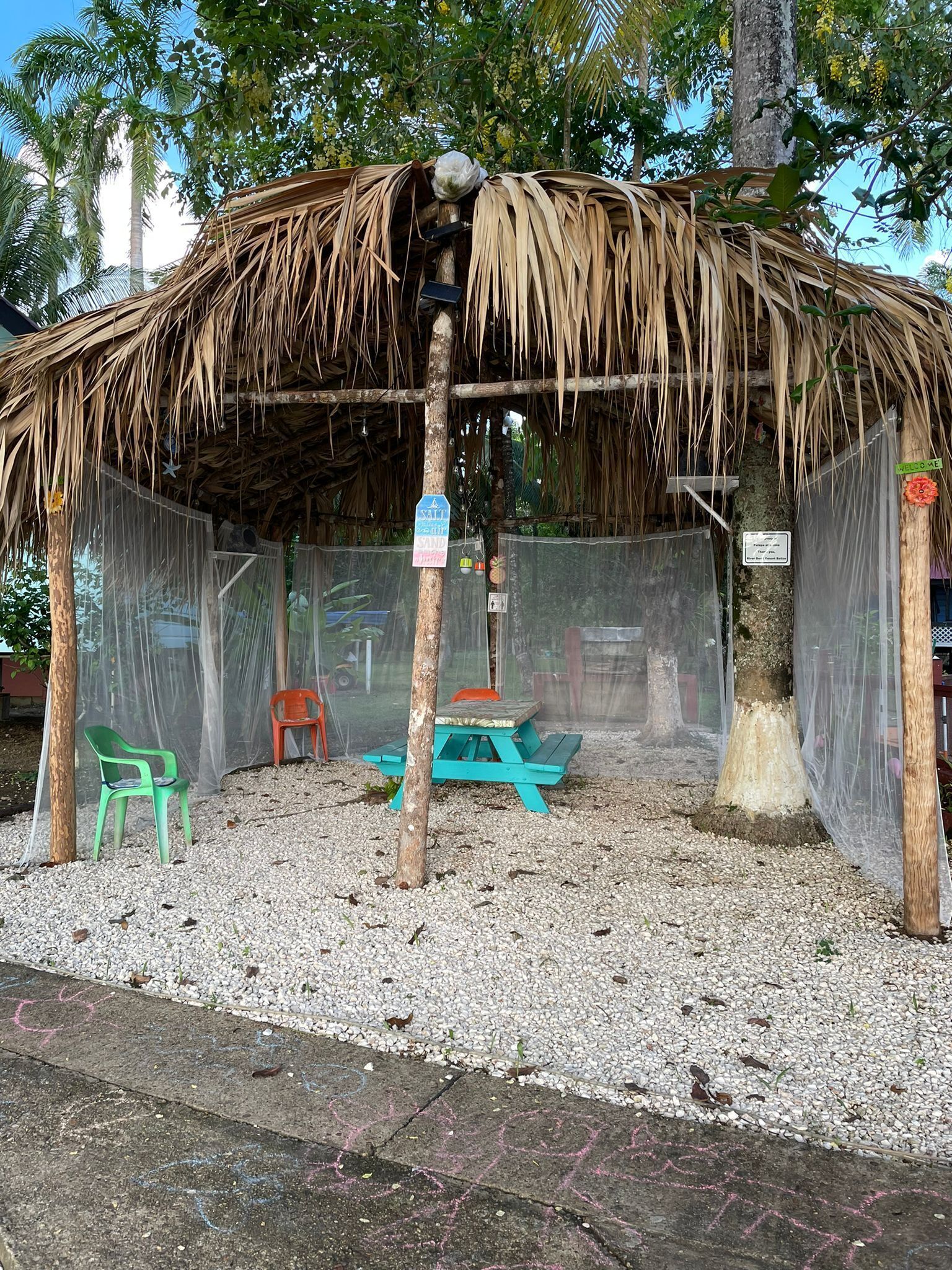 Une construction à toit de chaume, dotée d’une table de pique-nique et de chaises, entourée d’une clôture et d’arbres.