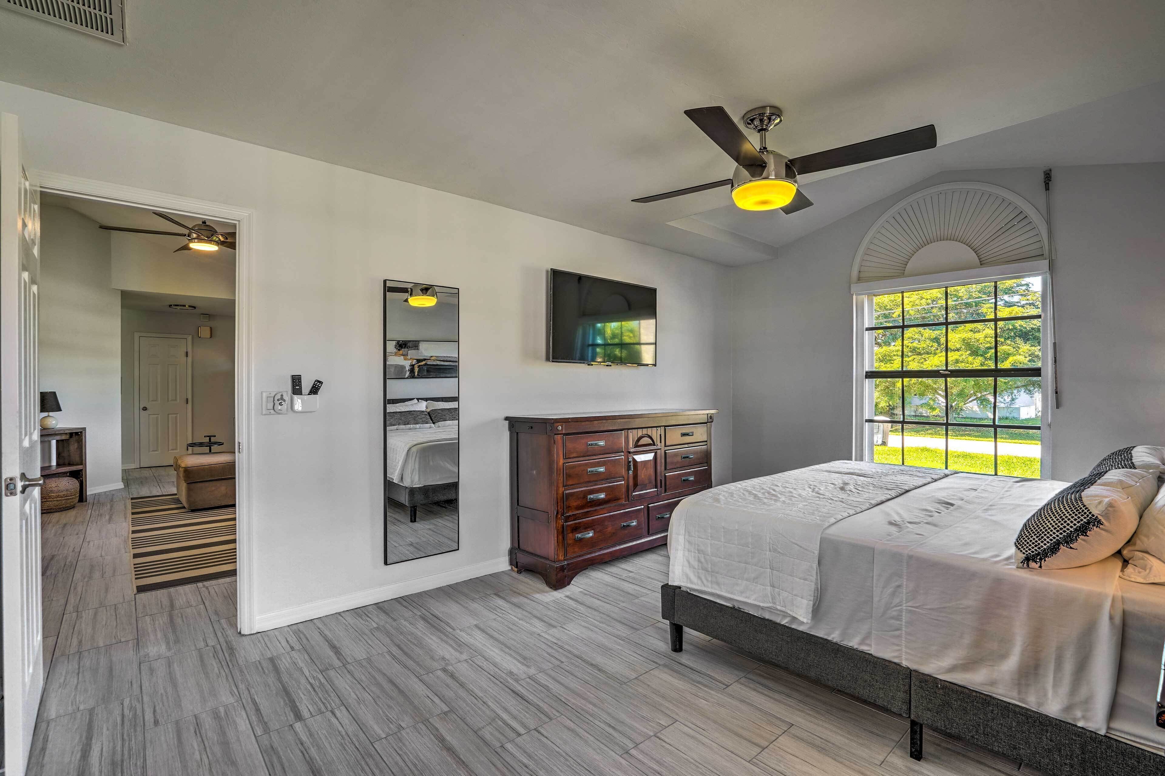 A bedroom with a bed, dresser, mirror, ceiling fan, and a window with a view of greenery.
