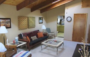 A cozy living room with a sofa, two armchairs, a coffee table, and a small table with a lamp. There is a door and a kitchen visible in the background.