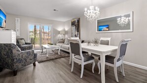 A modern living room with a dining area, featuring a chandelier, a large painting, and a television on a stand. - Spacious 2 bedroom condo (North Charleston)