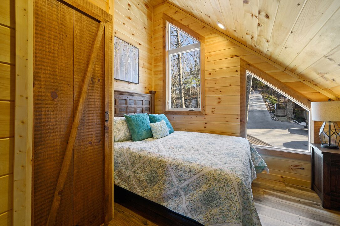 A bedroom with a wooden ceiling, a bed with patterned bedding, a window, and a view of a road outside.