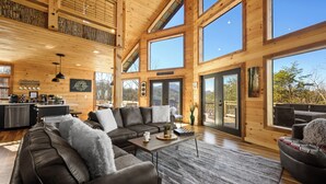 A spacious living area with a large sectional sofa, a coffee table, and a view of the outdoors through large windows. - BeautifulMtViewResortIndoorPool/Spa2CabinsPFNearIsland/Strip Game Room 16-18ppl (Pigeon Forge)