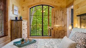 A wooden cabin bedroom with a large arched window, a bed with patterned bedding, a bedside table with a lamp, and a small plant on the windowsill. - BeautifulMtViewResortIndoorPool/Spa2CabinsPFNearIsland/Strip Game Room 16-18ppl (Pigeon Forge)