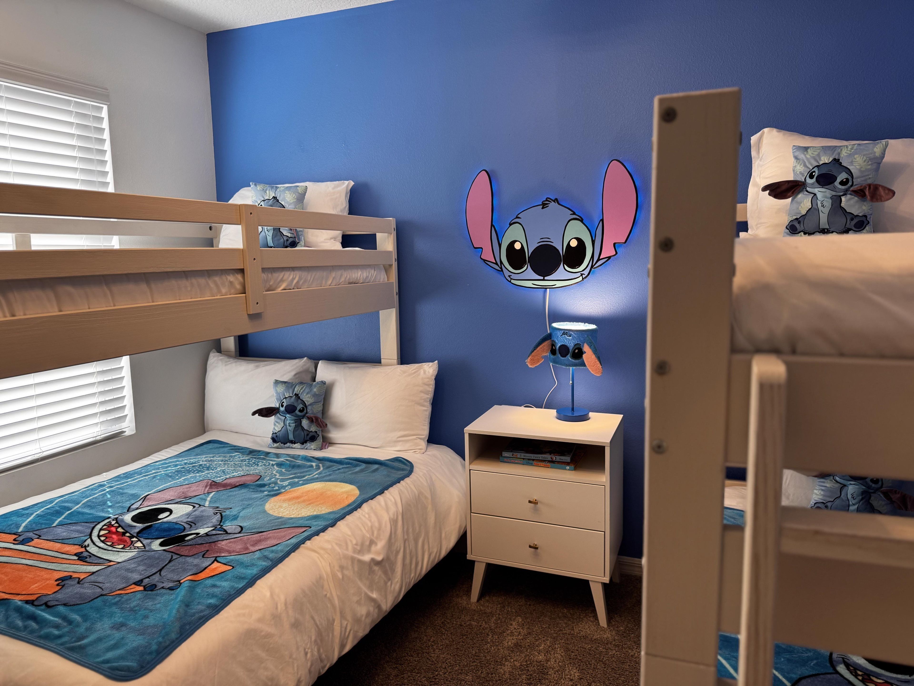 Une chambre d’enfant avec des lits superposés, une fresque murale représentant Stitch, une lampe de chevet et une table de chevet avec des livres.