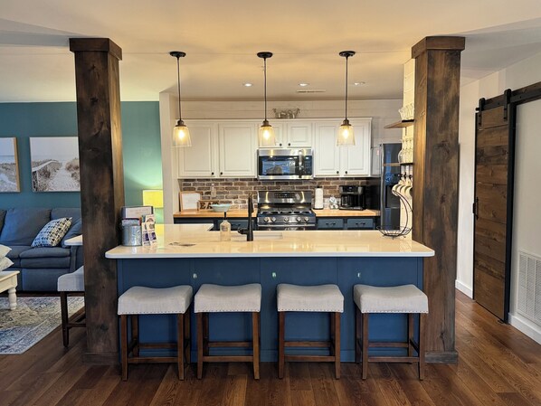A modern kitchen with a central island, bar stools, pendant lighting, and a view into the living area.