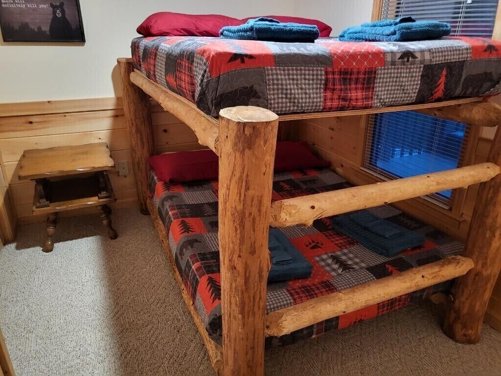 A bunk bed with a checkered blanket, a small wooden stool, and a framed picture on the wall.