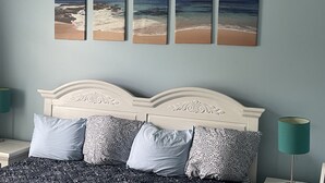 Un dormitorio con cama, almohadas, una mesita de noche, una lámpara y una pared con cuatro cuadros enmarcados.