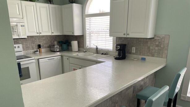 Una cocina moderna con armarios blancos, una isla grande con taburetes de bar, una cafetera y un ventanal con persianas.