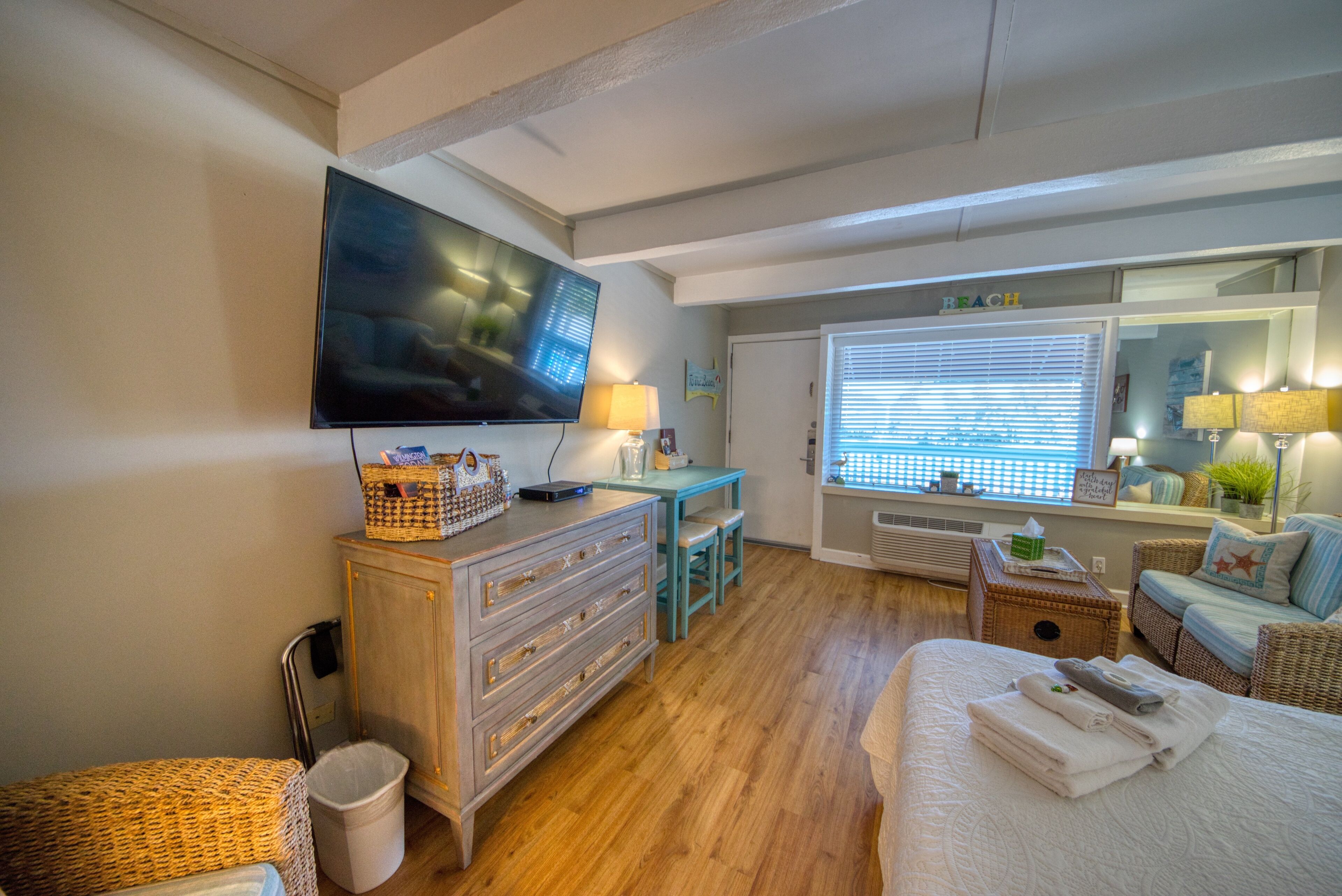Une chambre d’hôtel équipée d’une télévision, d’un bureau avec une lampe, d’un fauteuil en osier, d’un lit avec des serviettes et d’une fenêtre avec des stores.