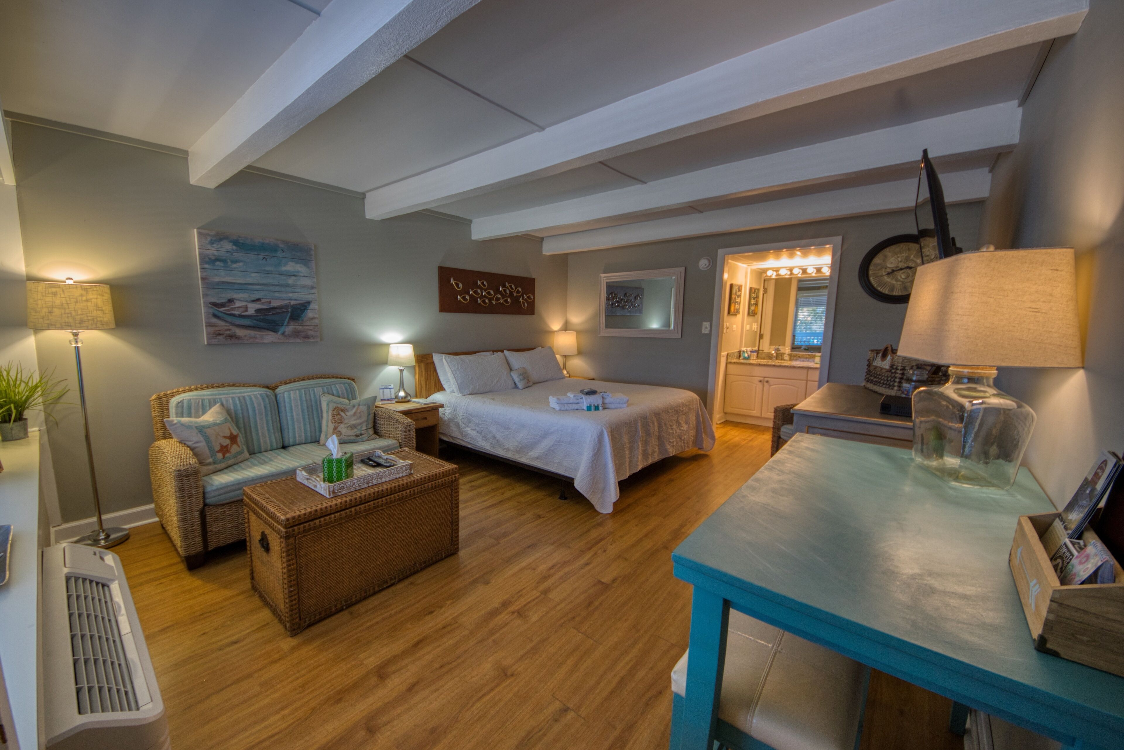 A cozy room with a bed, a sofa, a table, and a desk. There is a lamp, a clock, and a picture on the wall.
