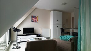 Quarto moderno e bem iluminado com televisão, secretária com computador, cadeira, sofá e radiador.