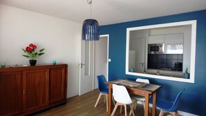 Zona pranzo con tavolo in legno e sedie blu, un armadietto con una pianta in vaso e una cucina visibile attraverso un'ampia finestra.