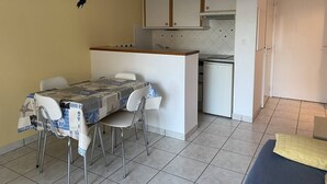 A compact kitchen with white cabinets, a small refrigerator, and a dining area with a table and chairs. - Location de Vacances - Pornichet-2 Pièces-005-60282 (PORNICHET)