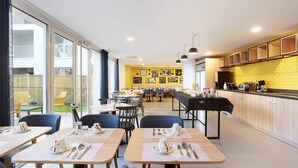 A modern dining area with tables set for breakfast, a kitchen area, and large windows with a view of the outside. - Studio Confortable avec Wi-Fi et Climatisation (Toulouse)