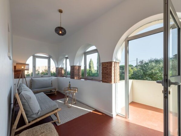 Un balcon spacieux offrant une vue sur la verdure, un coin salon avec un canapé et une petite table, ainsi qu’un luminaire suspendu.