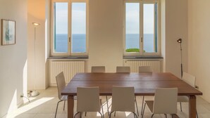 Ein Esszimmer mit Holztisch und sechs Stühlen, großen Fenstern mit Meerblick, einer Stehlampe und einem Heizkörper unter den Fenstern.