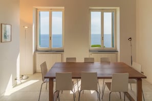 Comedor con mesa de madera y seis sillas, ventanas amplias con vista al mar, lámpara de pie y radiador debajo de las ventanas.