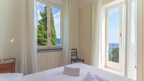 Ein Hotelzimmer mit Bett, Stuhl, Fenster mit Blick auf Bäume und Meer sowie einer Heizung.