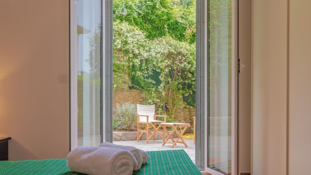 A room with a bed, a green patterned bedspread, a pair of rolled towels, and a view of an outdoor area with a chair and a table through a glass door.