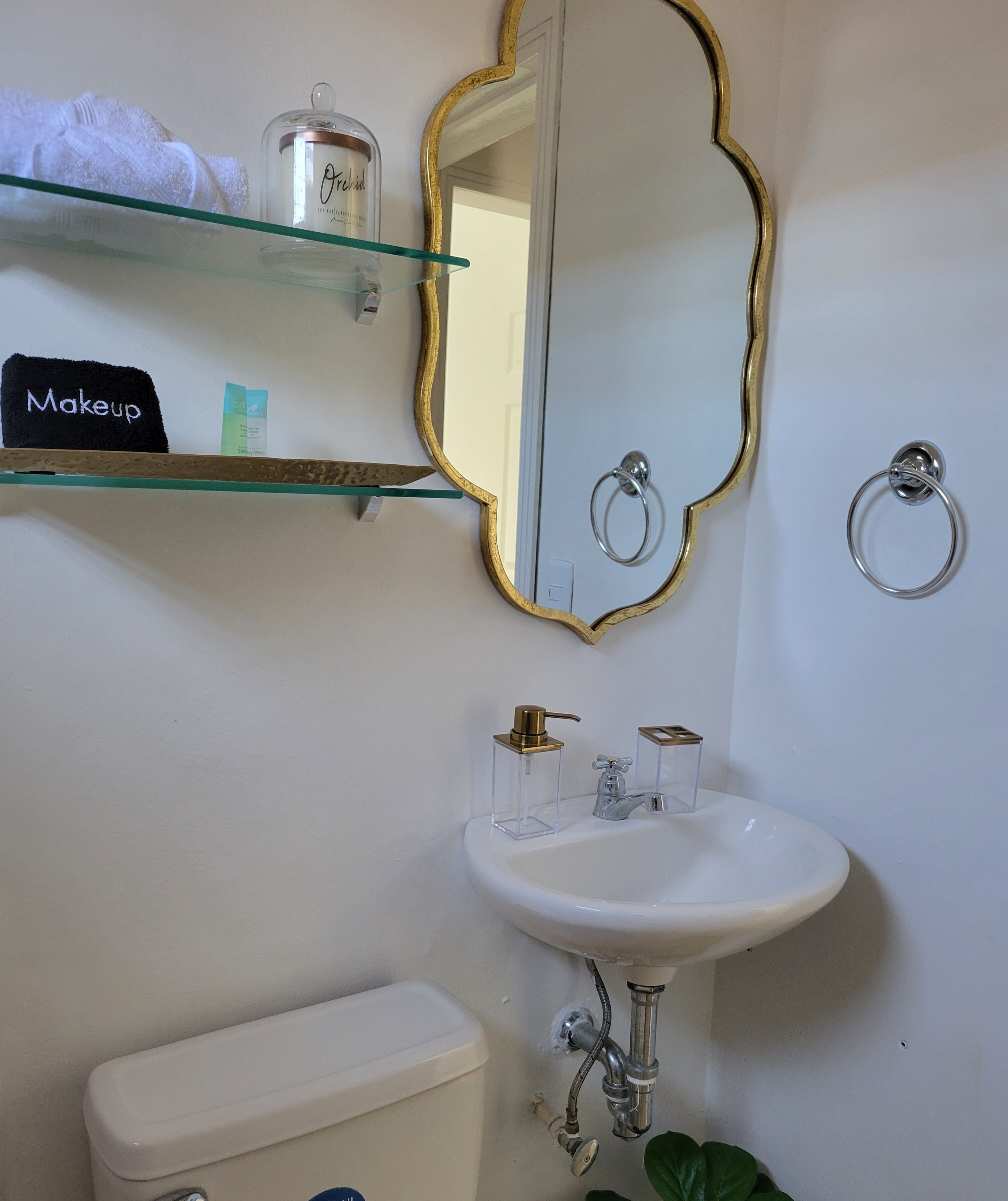 A bathroom with a white sink, gold-framed mirror, and a glass shelf with toiletries.