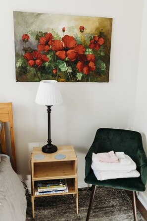 A bedroom with a bed, a green armchair, a wooden nightstand with a lamp, and a painting of red flowers on the wall.