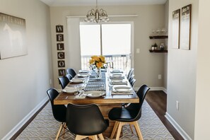 A dining area with a long wooden table set for six, surrounded by black chairs, with a 'WELCOME' sign on the wall and a chandelier above.
