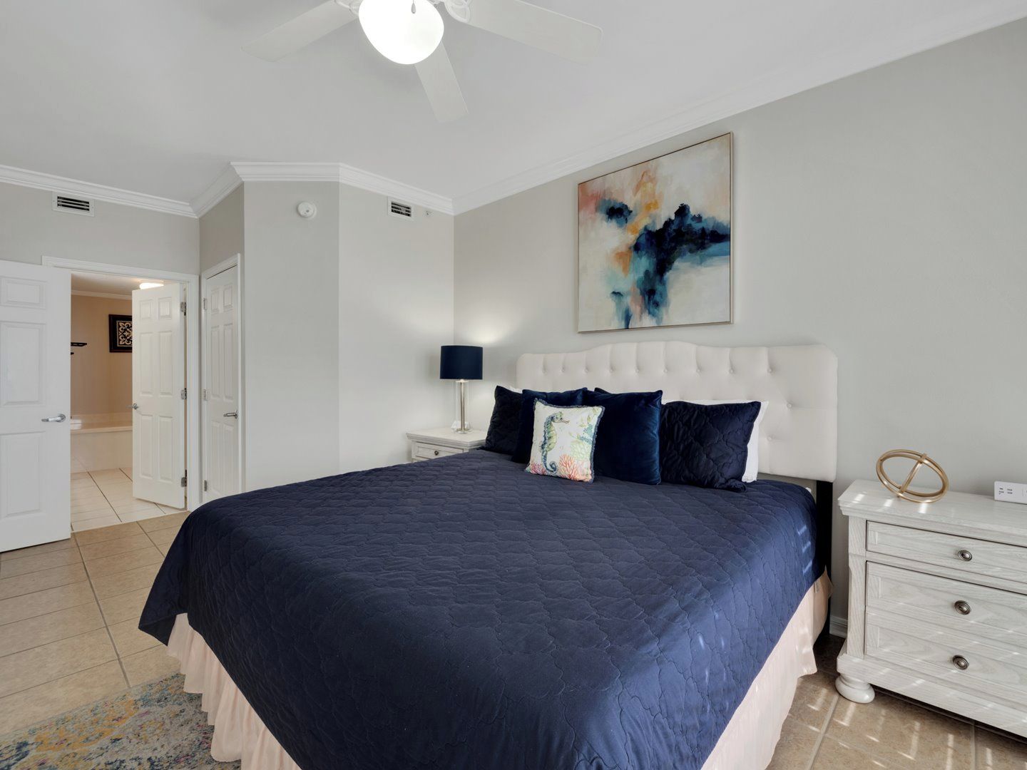 A bedroom with a large bed, white nightstands, a ceiling fan, and a painting on the wall.