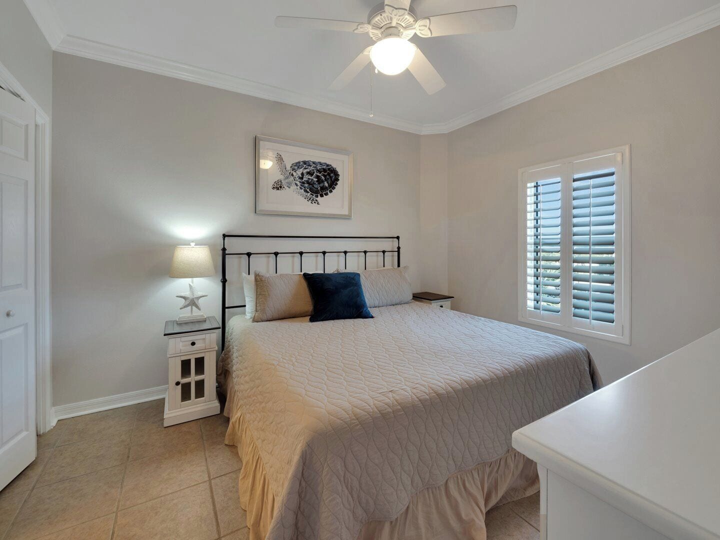 A bedroom with a bed, a nightstand, a ceiling fan, a window with blinds, and a framed picture on the wall.