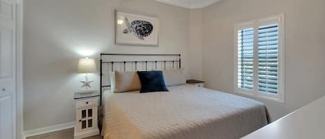 Une chambre à coucher comprenant un lit, une table de chevet, un ventilateur de plafond, une fenêtre avec des stores et un tableau encadré accroché au mur.