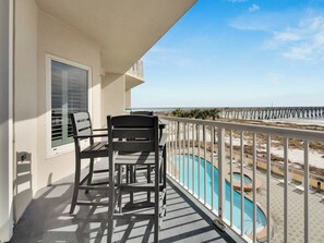A balcony with two chairs and a table, overlooking a swimming pool and a beach with a pier.