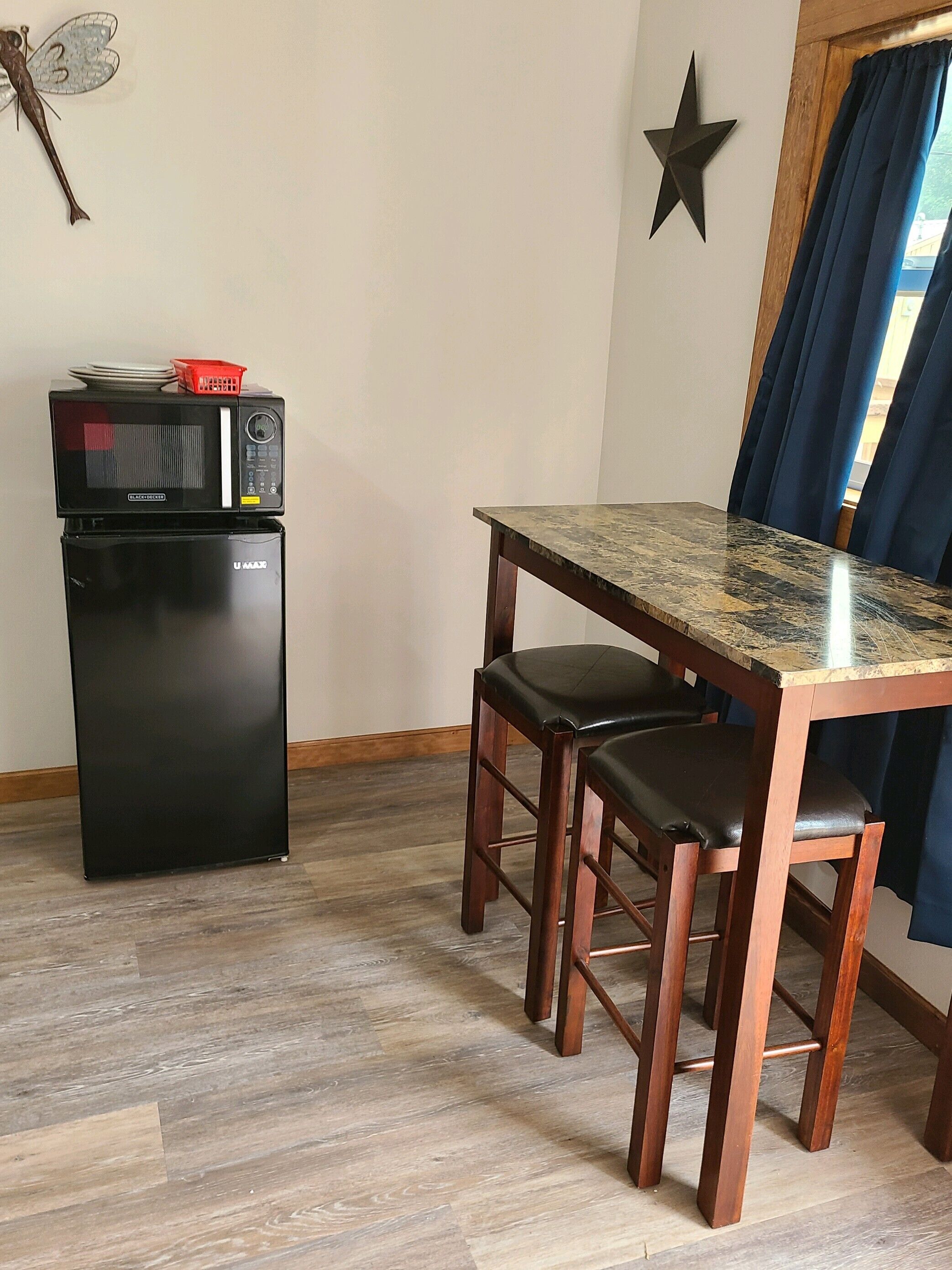 A compact kitchenette with a microwave and refrigerator, a wooden dining table with two chairs, a star decoration on the wall, and a window with blue curtains.