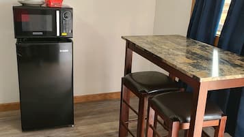 A compact kitchenette with a microwave and refrigerator, a wooden dining table with two chairs, a star decoration on the wall, and a window with blue curtains.