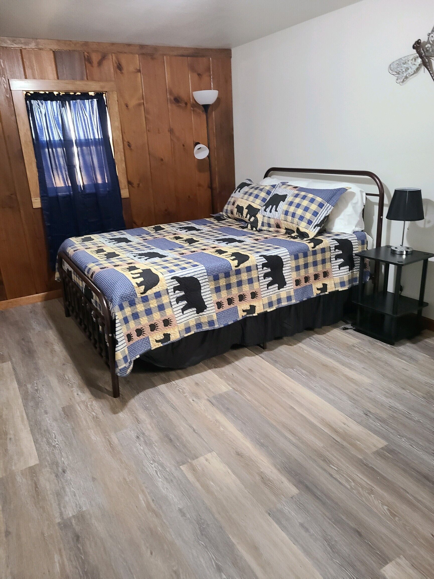 Un dormitorio con cama, mesita de noche, lámpara, ventana con cortinas y paredes de madera.