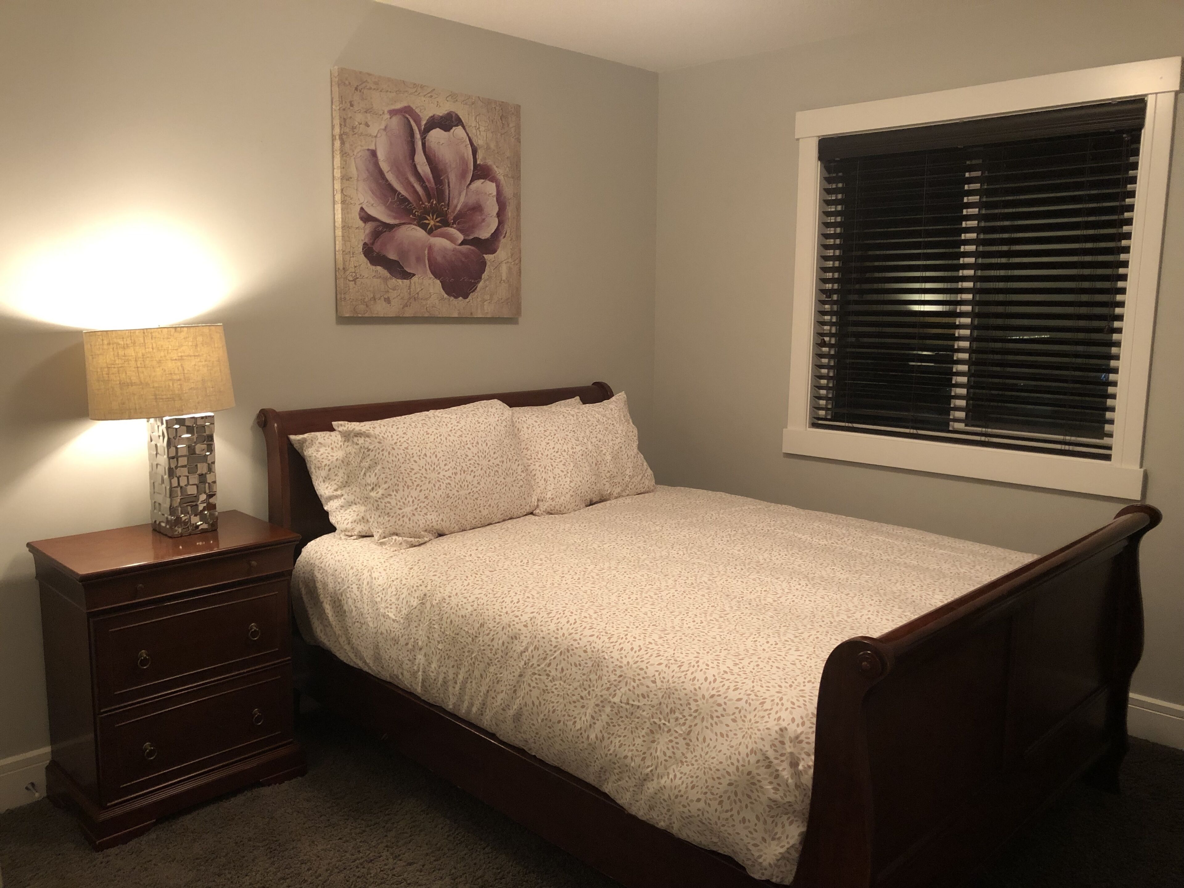 Una camera da letto con un letto di legno, un comodino con una lampada, una cassettiera, un quadro con fiori sulla parete e una finestra con persiane.