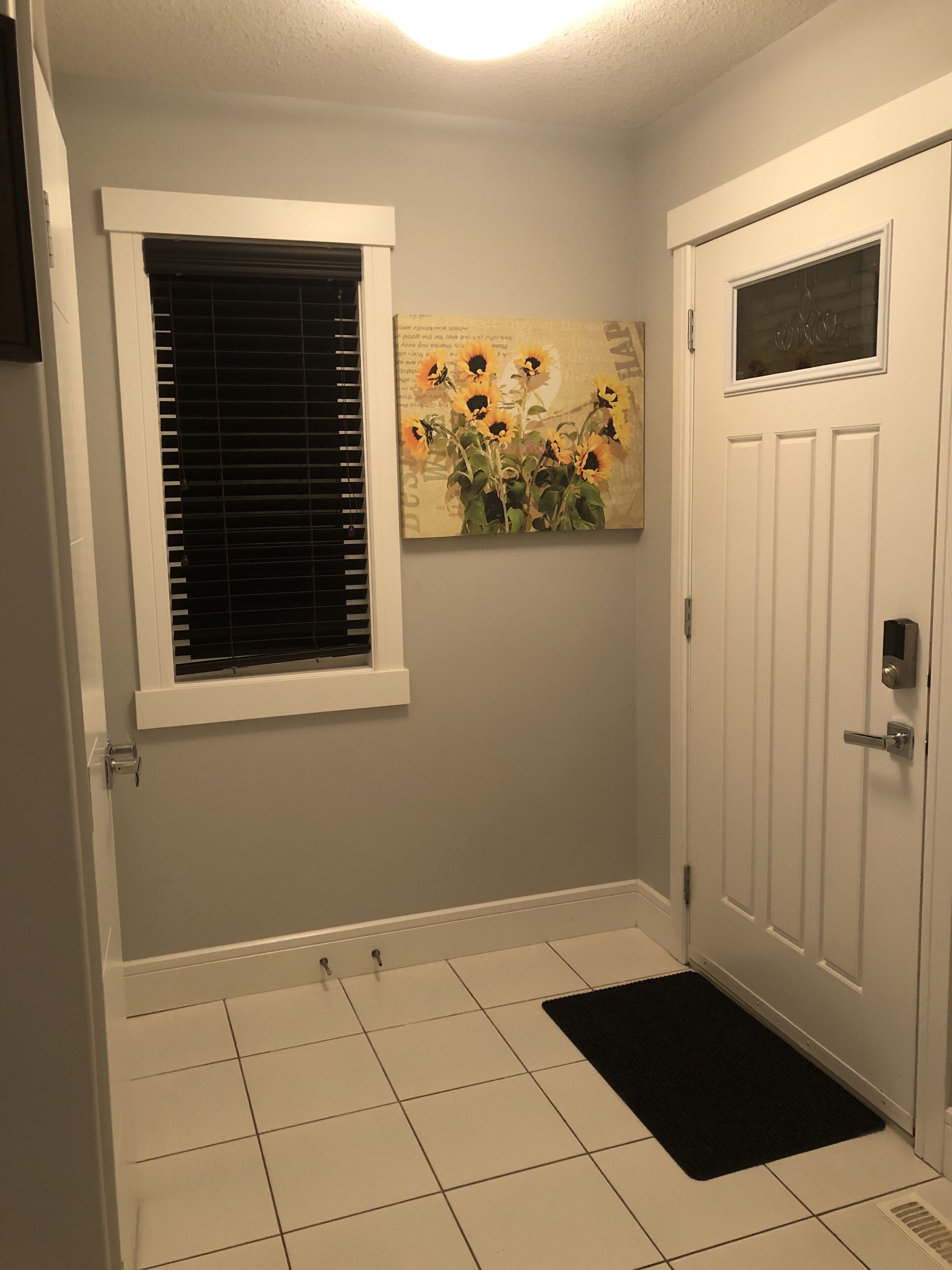 A room with a white door, a window with blinds, a black doormat, and a painting of sunflowers on the wall.