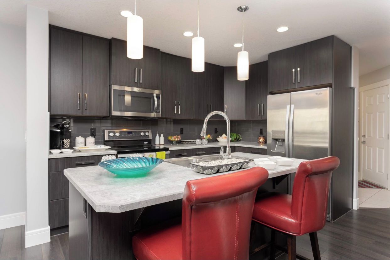 A modern kitchen with dark cabinets, stainless steel appliances, a marble countertop, and red leather chairs.