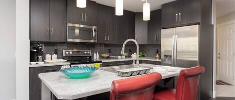 A modern kitchen with dark cabinets, stainless steel appliances, a marble countertop, and red leather chairs.